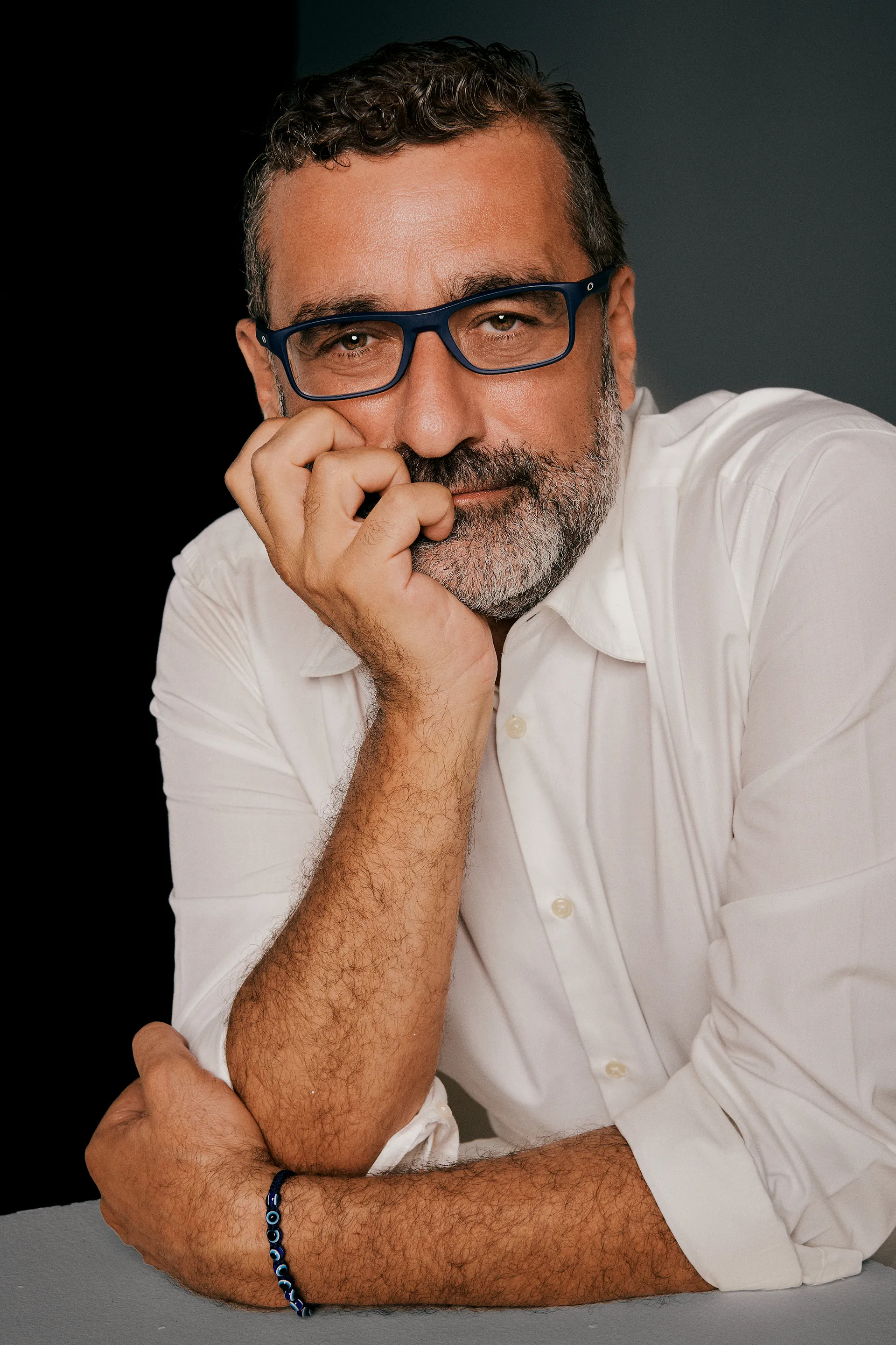 Middle-aged man with curly hair and beard wearing glasses and white shirt resting head on hand.