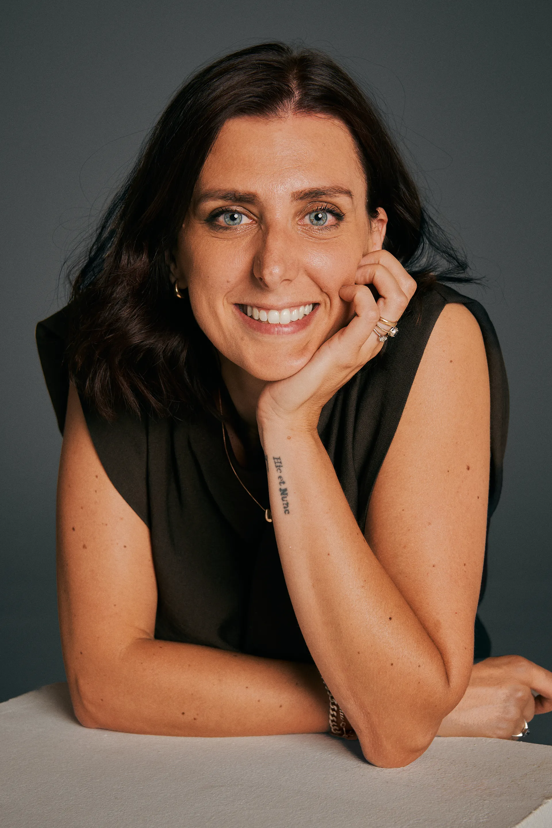 Smiling woman with dark hair resting her chin on her hand against a dark gray background.