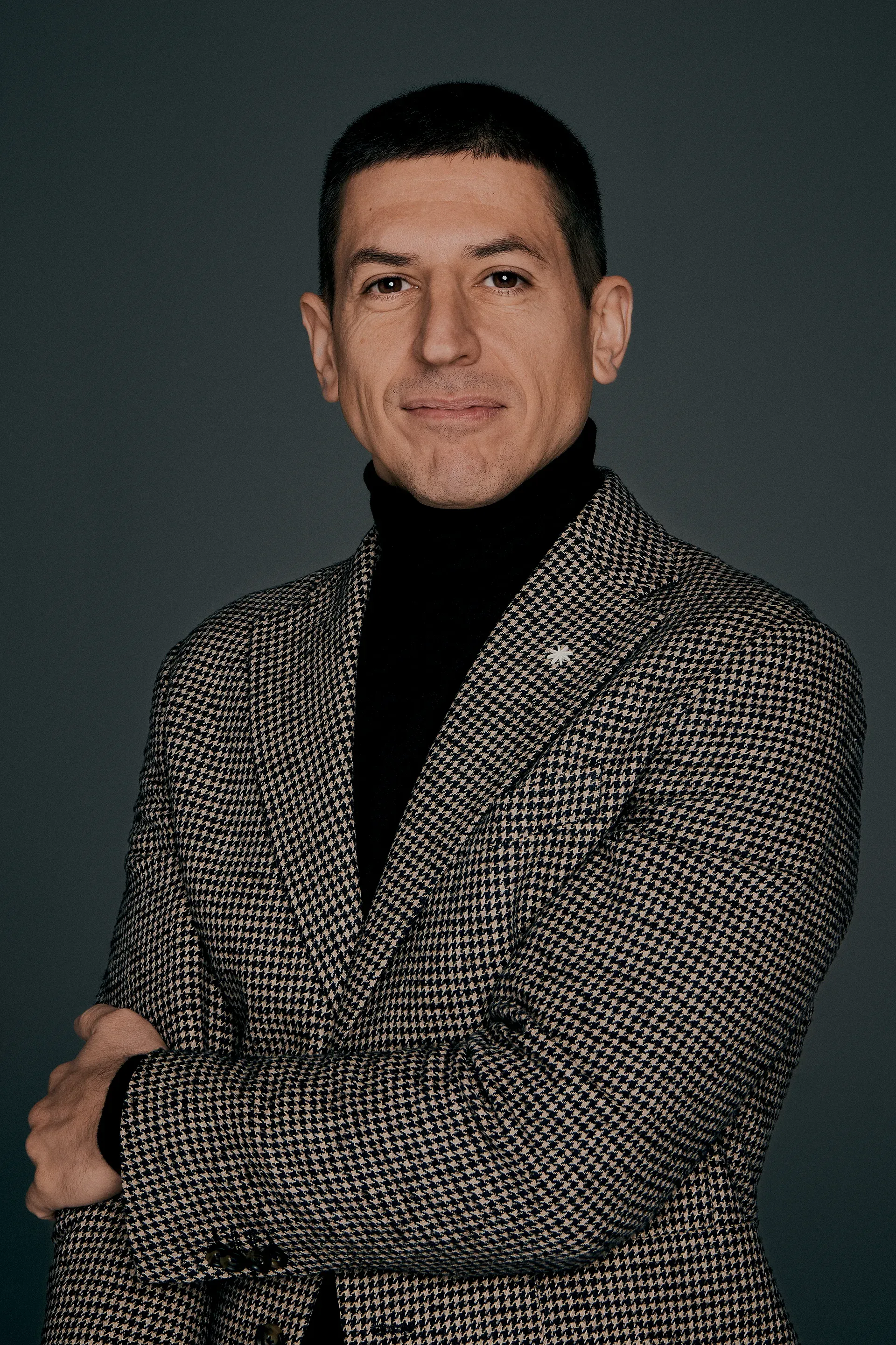 Man with short black hair wearing a houndstooth blazer and black turtleneck against dark background.