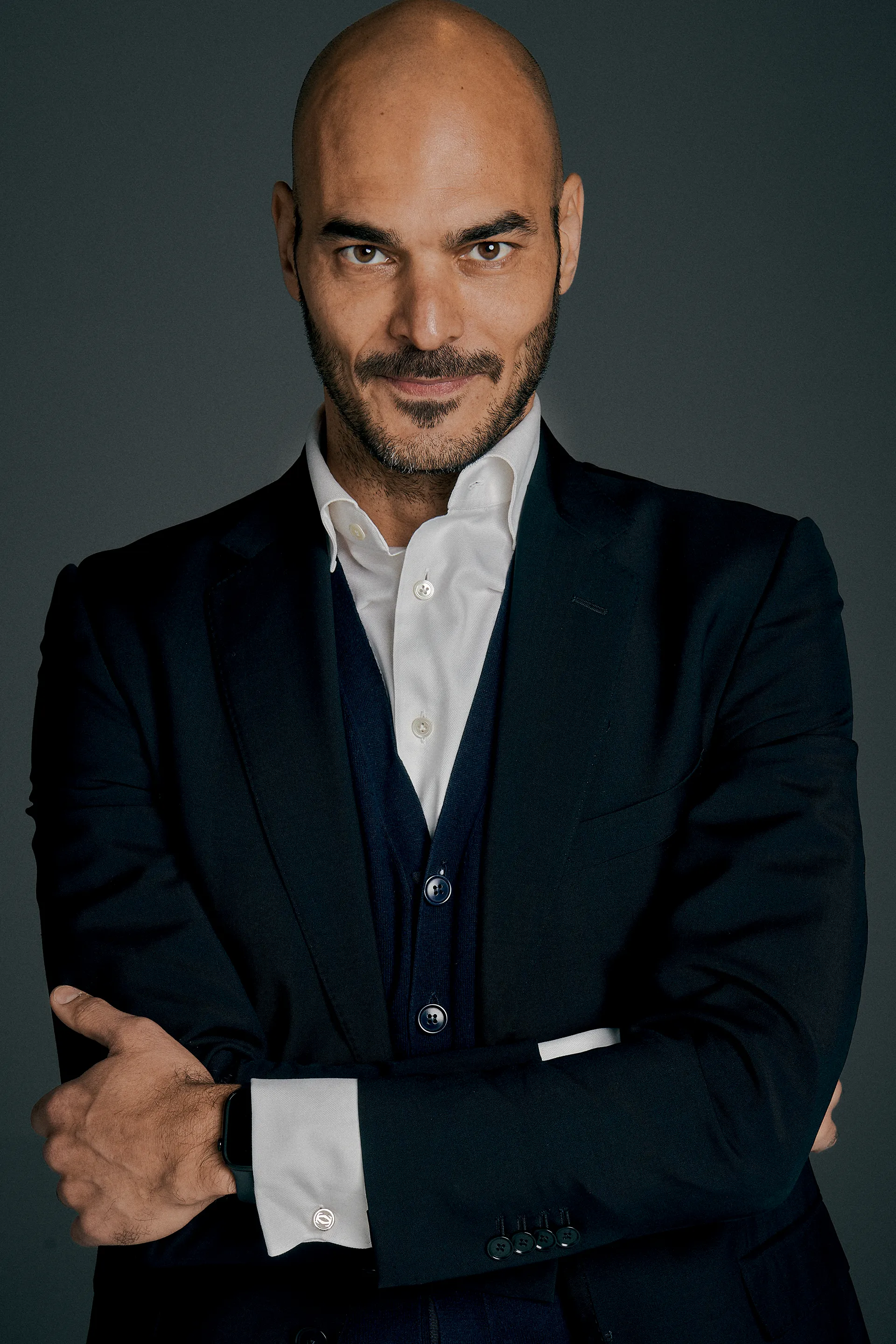 Confident bald man with trimmed beard wearing dark suit and white shirt with arms crossed.