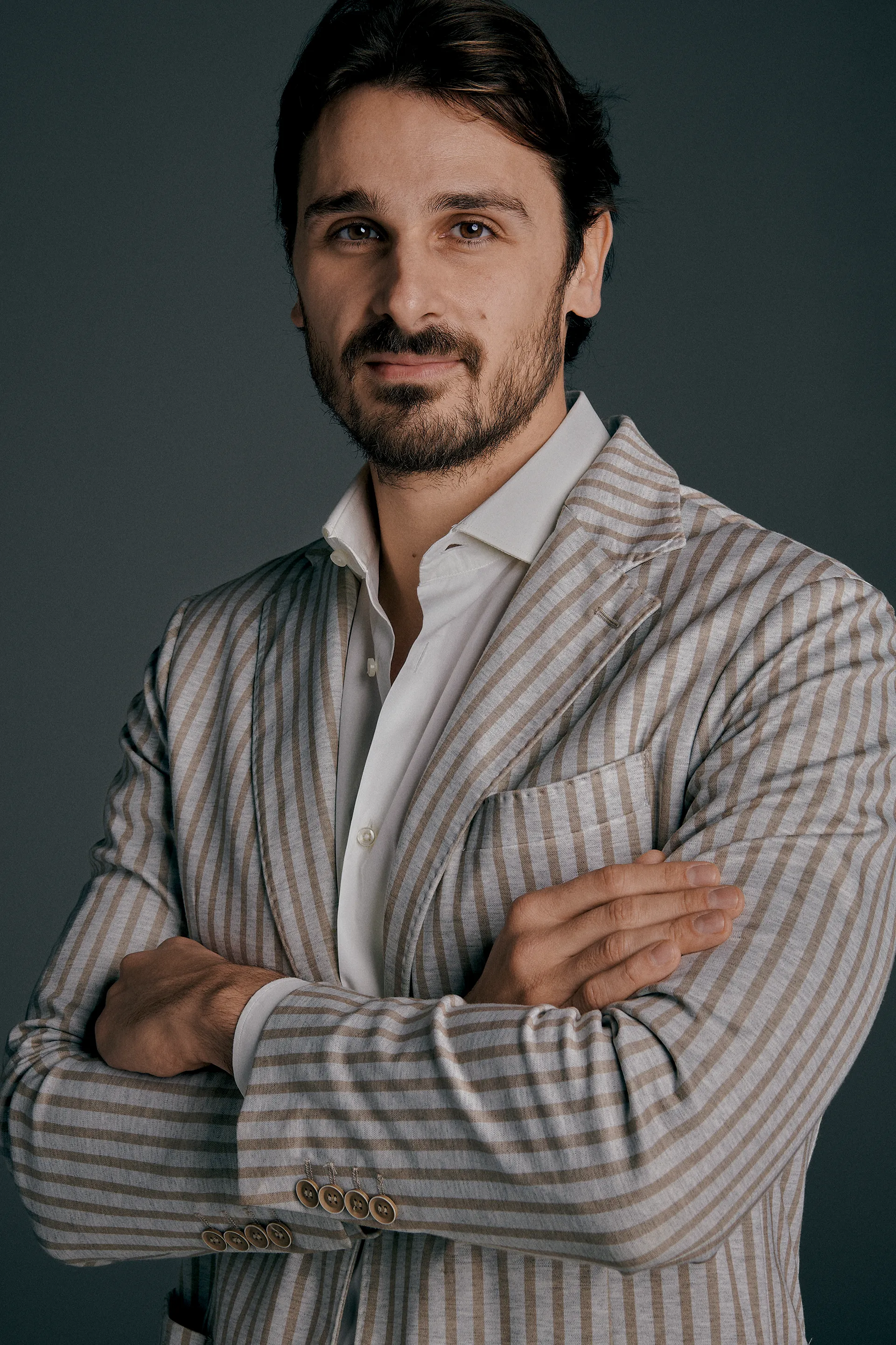 Man with dark hair and beard wearing striped blazer and white shirt against a dark background.