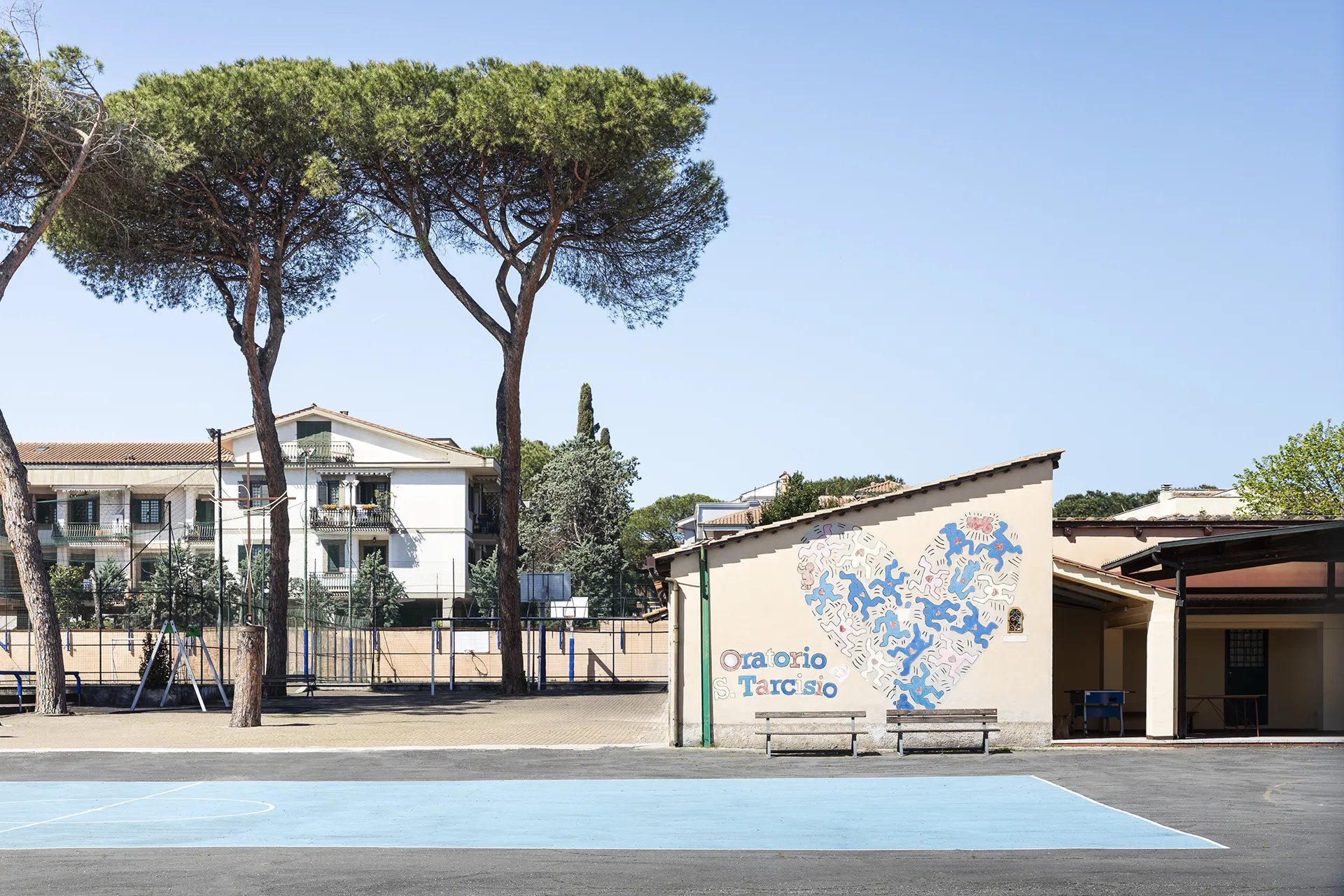 Outdoor basketball court with tall pine trees and a building wall mural of blue human figures in a heart shape.