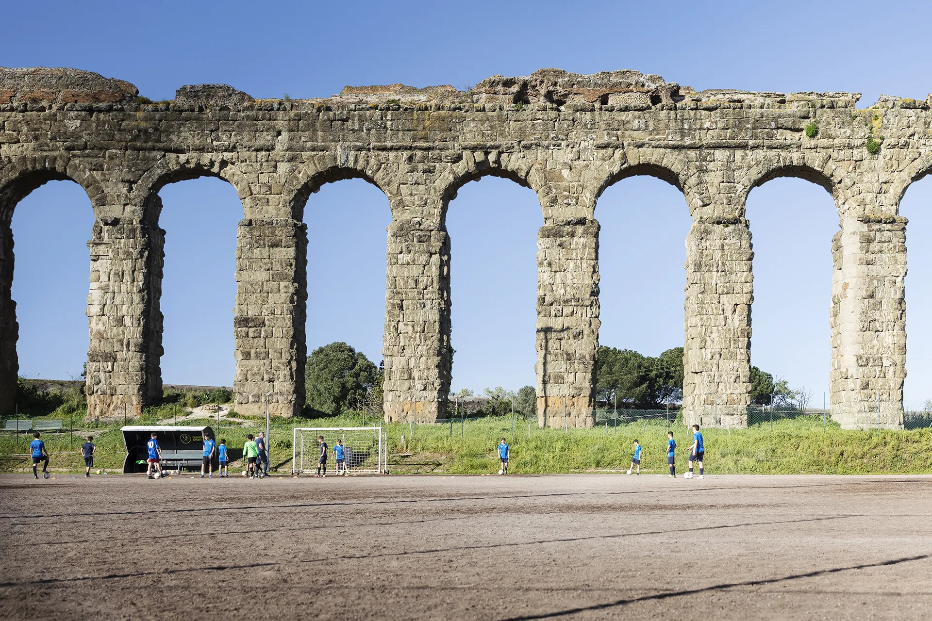 Children playing soccer on a field in front of a large ancient stone aqueduct with arches.