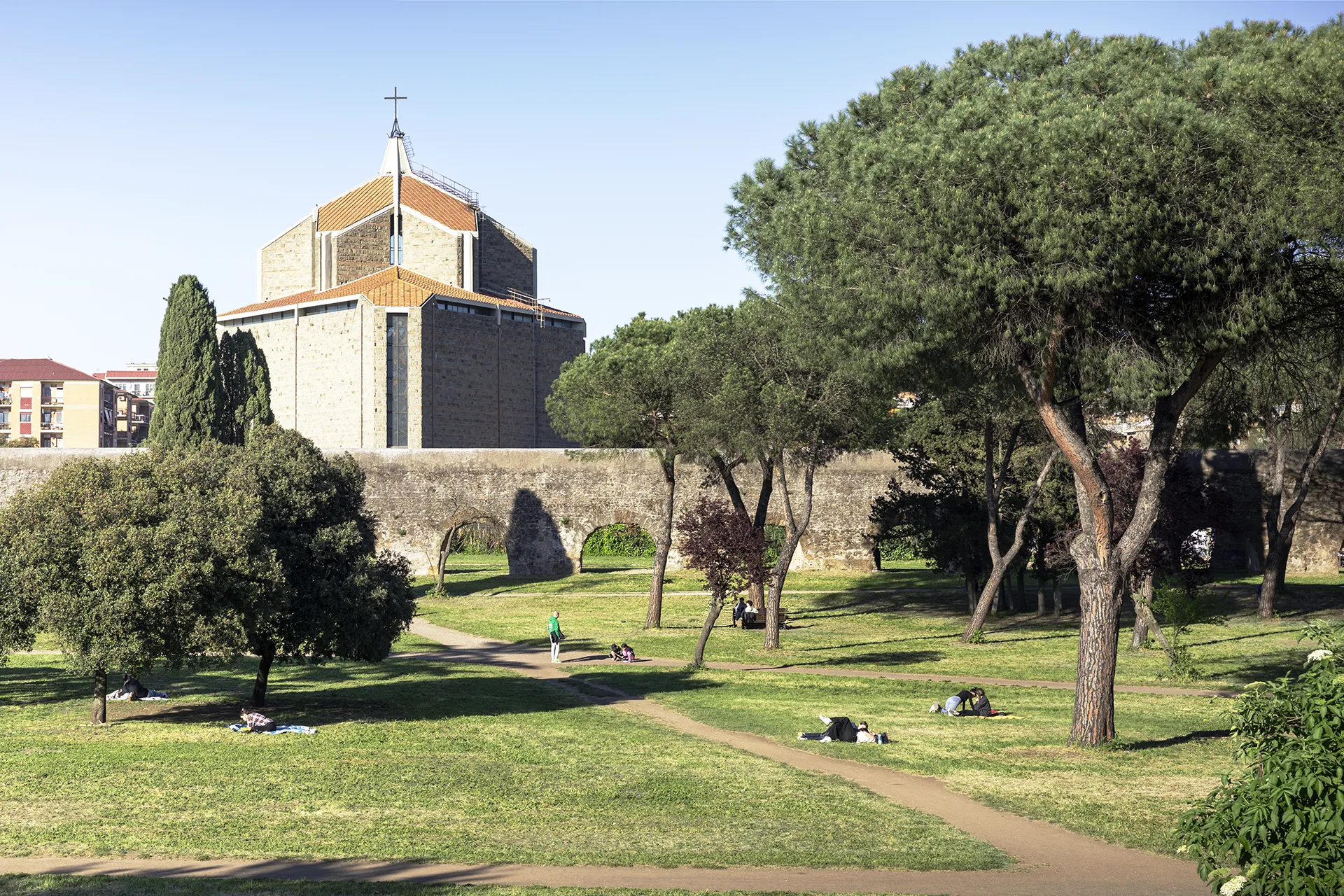 People relaxing on grass and benches near trees with a church and stone wall in the background.