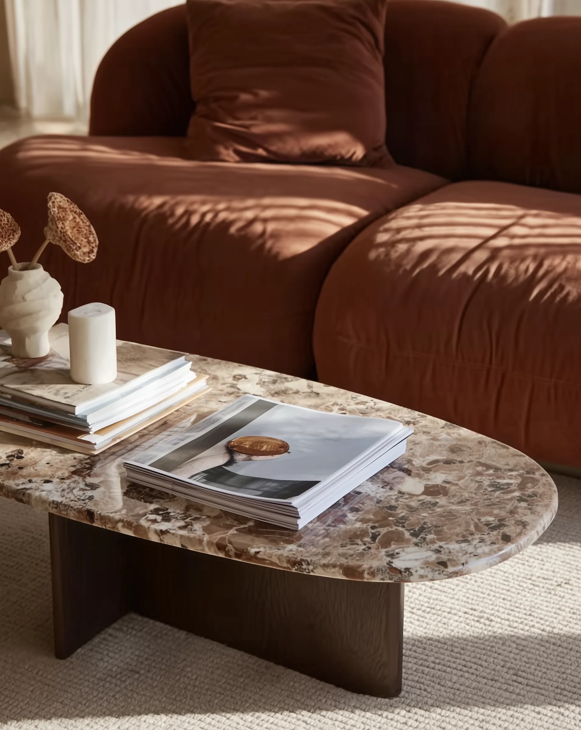 Oval marble coffee table with books, a candle, and dried flowers in a beige vase in front of a brown sofa.